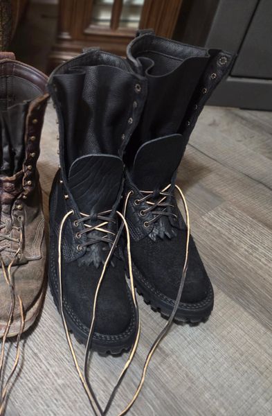 A pair of black leather boots with laces and decorative studs sitting on a wooden floor next to brown boots