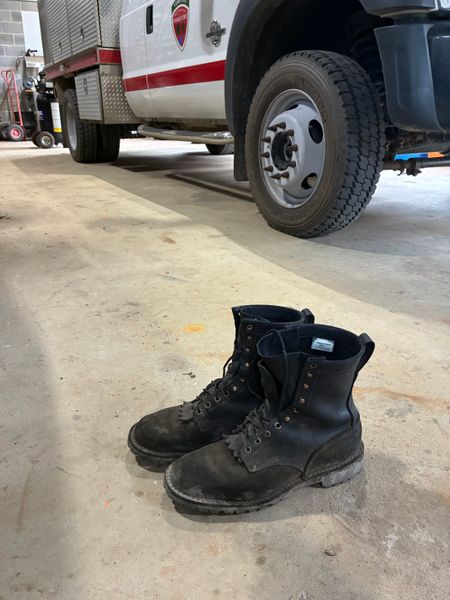 A pair of black work boots sits on a concrete floor next to a fire truck in what appears to be a fire station garage.