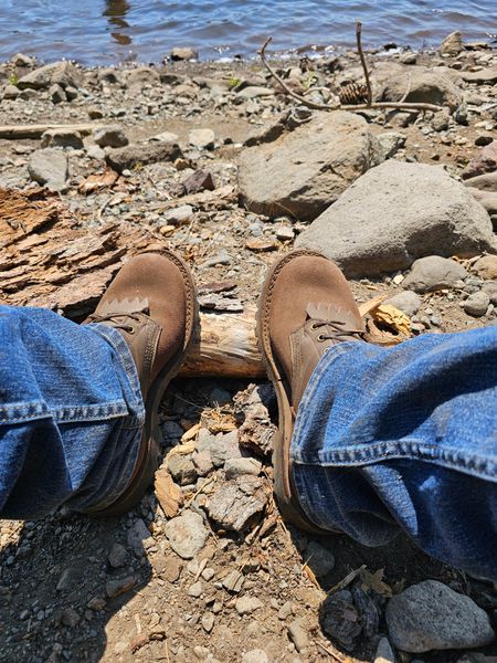 Person wearing jeans and brown boots sitting on rocky shoreline near water