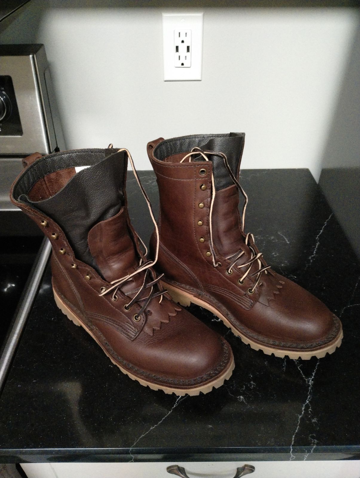 A pair of brown leather work boots with laces sitting on a dark marble countertop in a kitchen