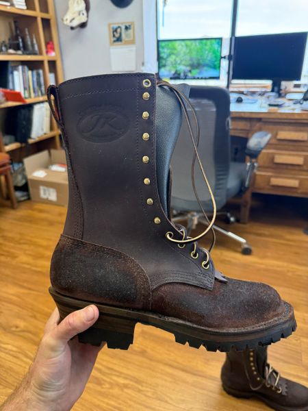 A person holding a tall black leather boot with laces and brass eyelets in an office setting with wooden floors, bookshelves, and a desk with computer monitors in the background.