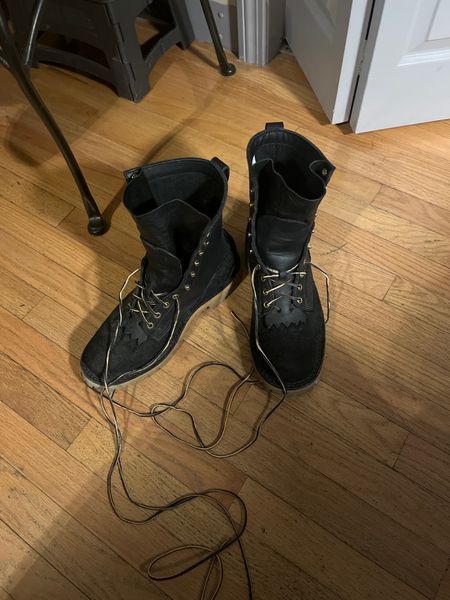 A pair of black leather boots with long laces sitting on a wooden floor next to chair legs
