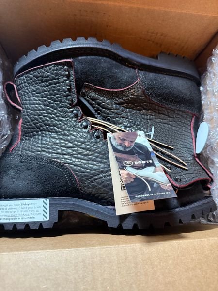 Black leather work boots with red stitching in a cardboard box with product tags attached