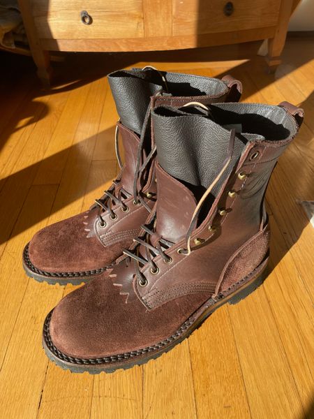 A pair of brown leather work boots with laces sitting on a wooden floor next to wooden furniture