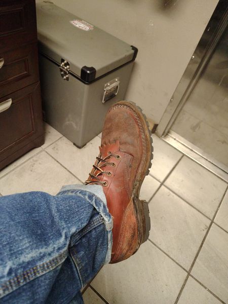 Person wearing worn brown leather work boots and jeans sitting in a bathroom with tiled floor and storage container visible