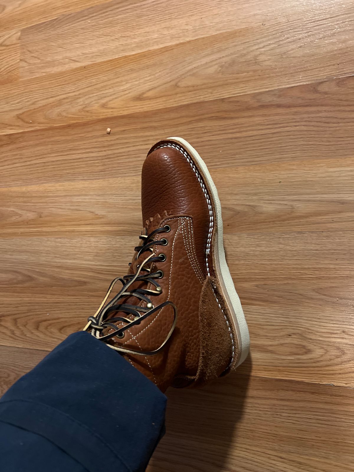 A brown leather lace-up shoe with white stitching on a wooden floor, worn with blue pants