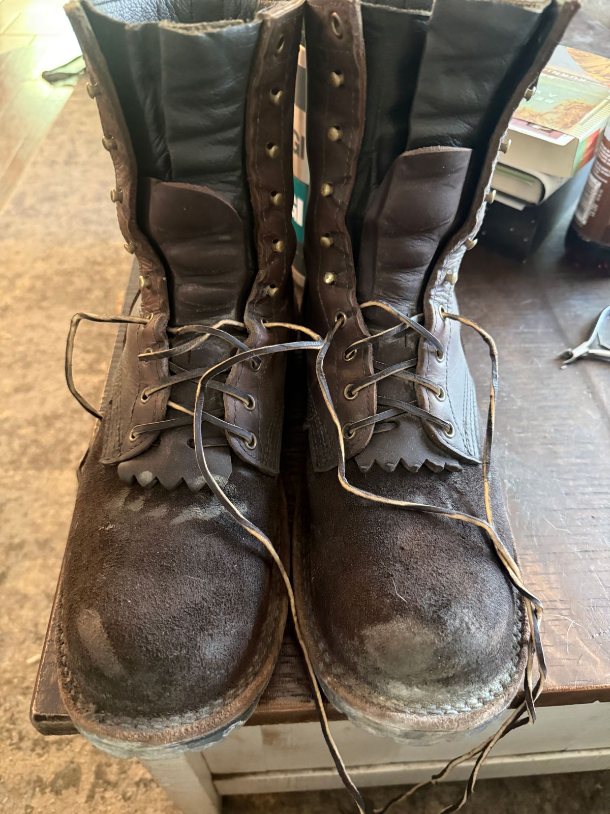 A pair of worn brown leather work boots with laces and metal eyelets sitting on a wooden surface