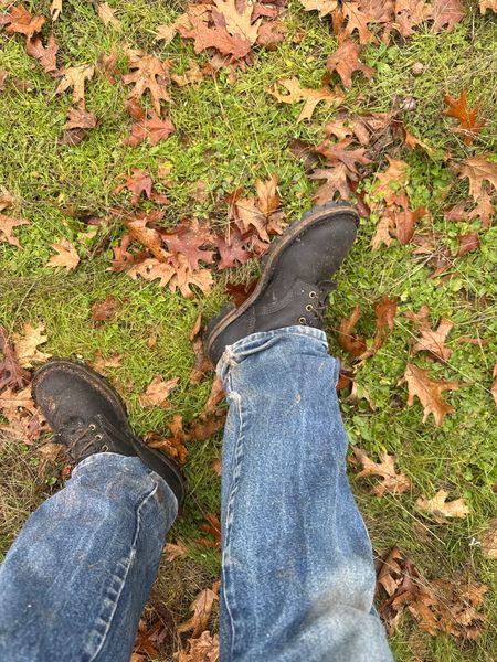 Person's legs in jeans and dark boots standing on grass covered with fallen autumn leaves