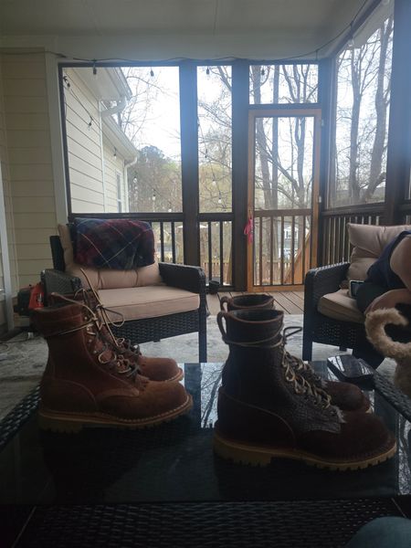 A screened porch with wicker furniture and cushions, featuring two pairs of boots on a coffee table in the foreground, with bare trees visible through the screens in the background