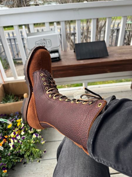 Person wearing brown leather lace-up boots with their feet up on a white deck railing, with flowers and outdoor furniture visible in the background