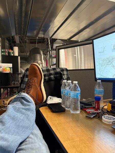 Person with feet up on desk in what appears to be a mobile office or RV workspace, with water bottles, computer monitor showing technical diagrams, and various equipment visible