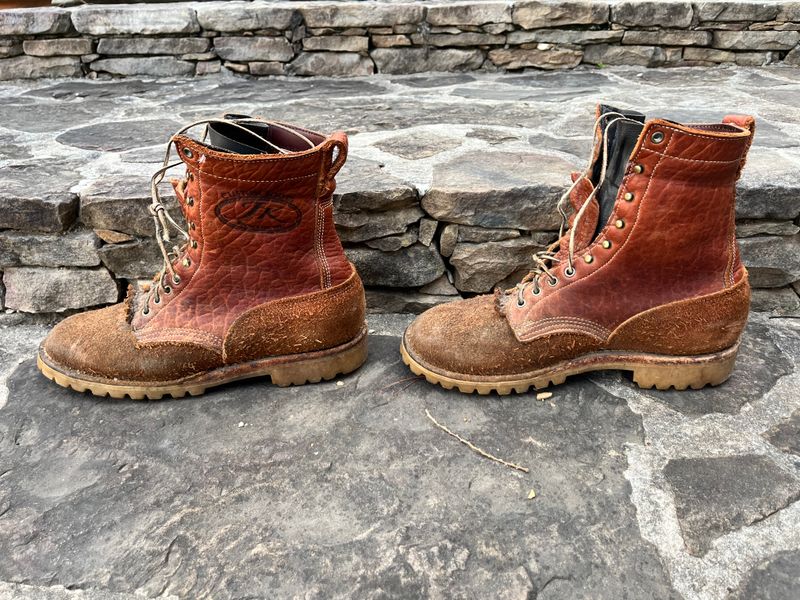 A pair of brown leather work boots with laces sitting on stone pavement next to a stone wall