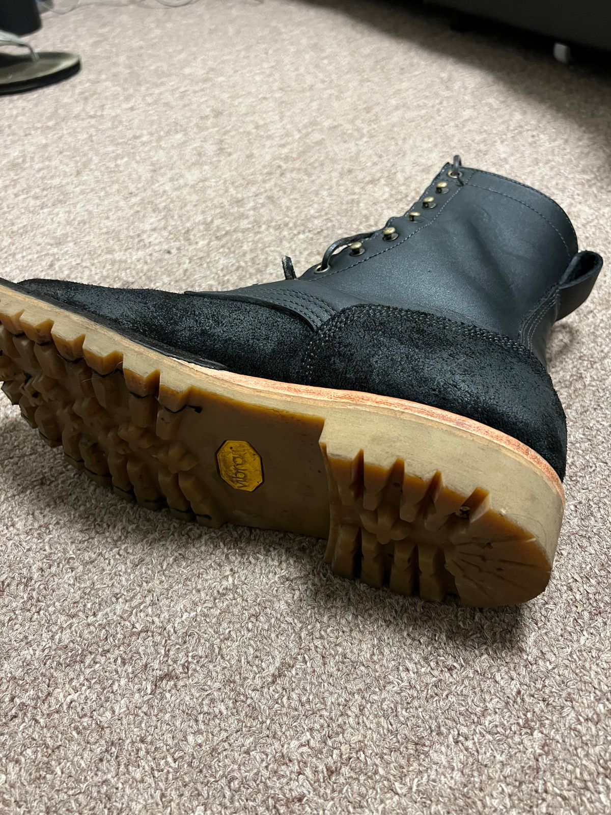 A black leather boot with laces lying on its side on carpet, showing the thick treaded sole with yellow Vibram logo