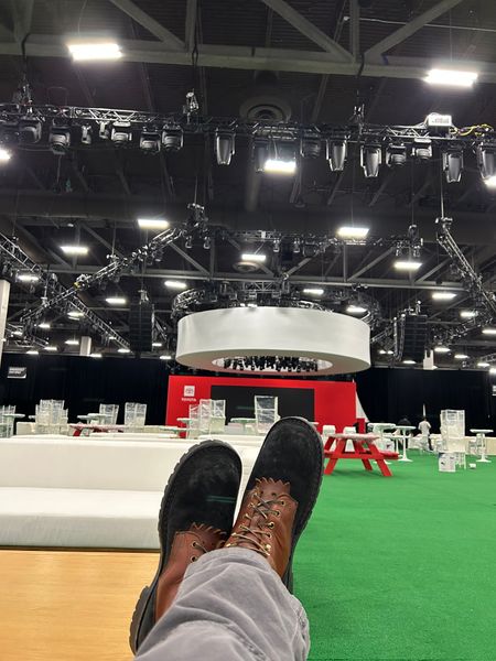 Person's feet in brown and black boots resting on a ledge overlooking a television studio set with green carpet, lighting equipment, and production infrastructure visible in the background