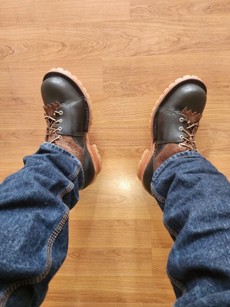 Person wearing dark leather boots with brown laces and blue jeans standing on wooden floor