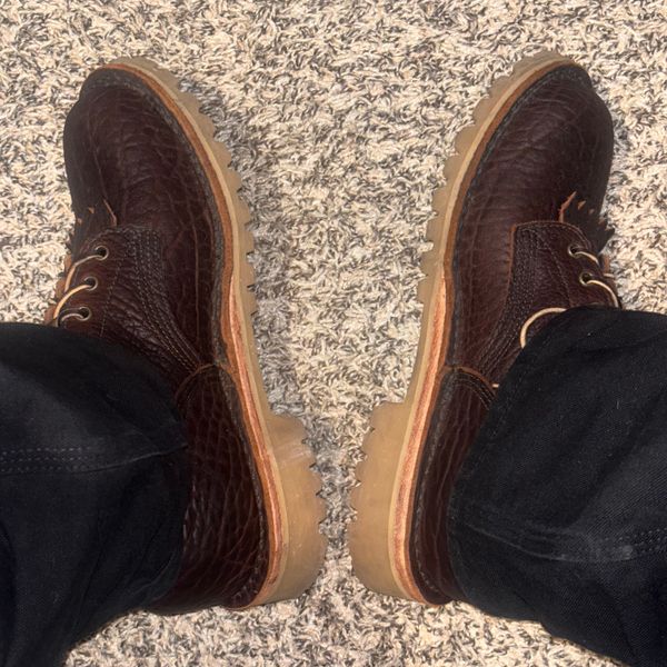 A pair of brown leather shoes with decorative fringe detail on a textured carpet, worn with dark pants