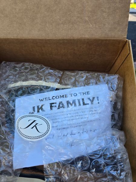 A cardboard shipping box containing bubble wrap packaging and a welcome card with a circular JK logo