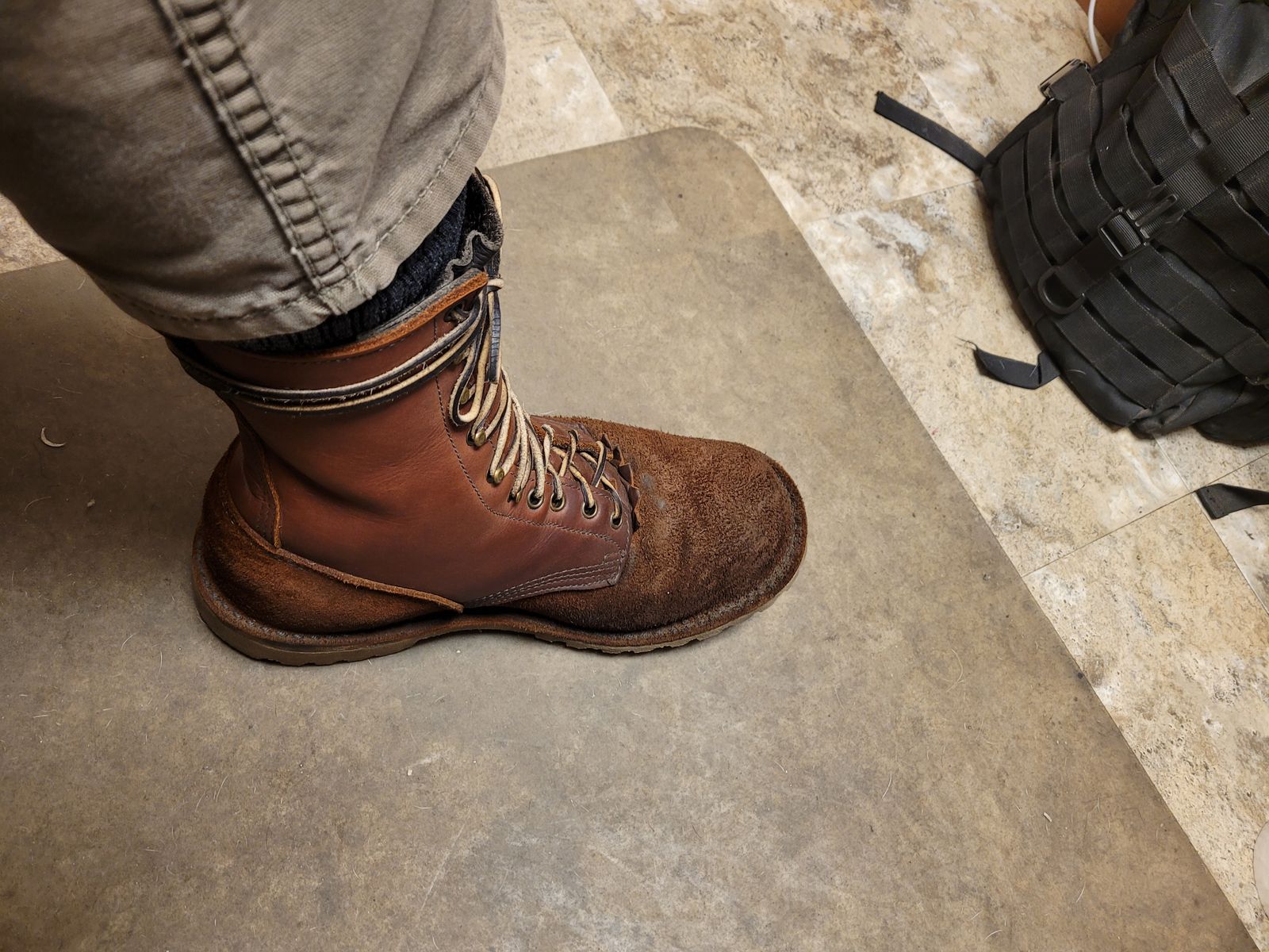 A person wearing brown leather work boots standing on a concrete floor next to military gear and equipment
