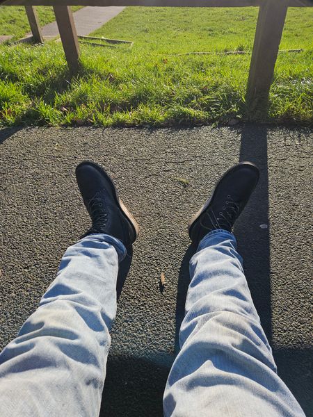 Person's legs and feet in jeans and dark sneakers sitting on asphalt path with green grass and fence visible in background