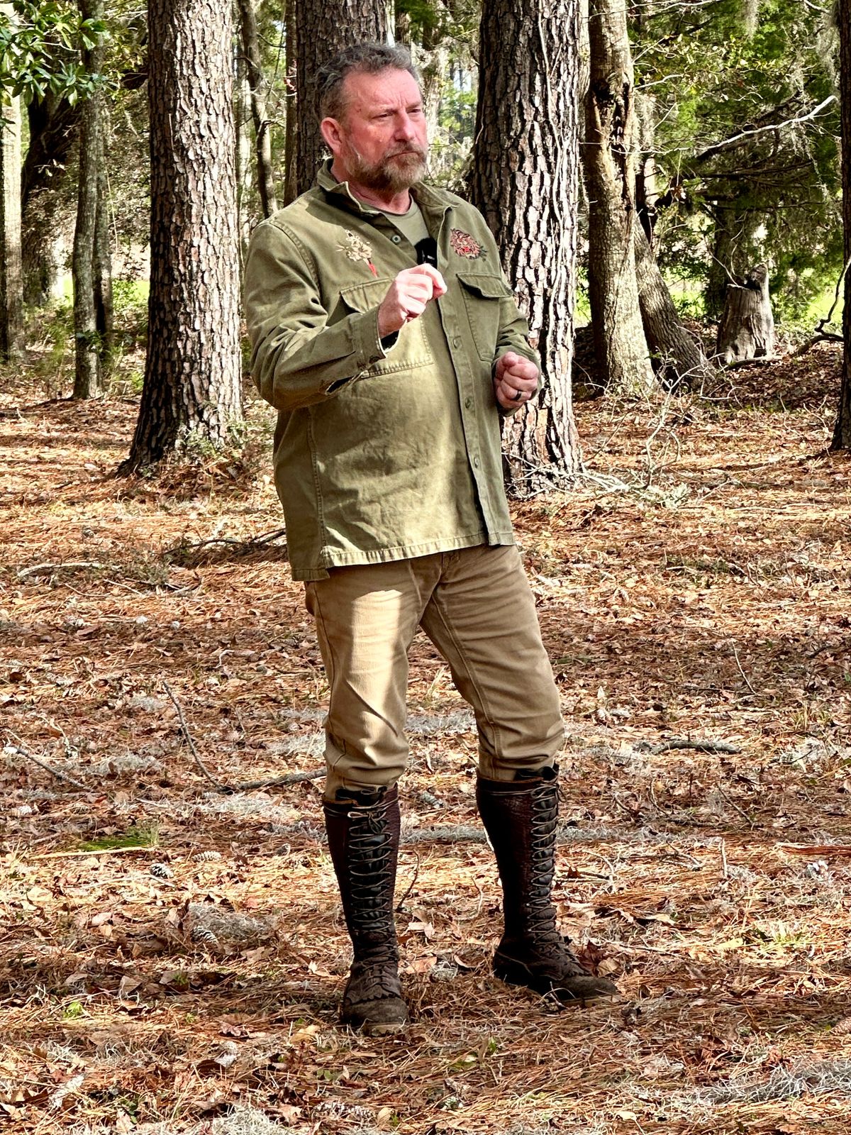 A man in outdoor clothing stands in a wooded area holding a white cup, wearing boots and earth-tone garments suitable for hiking or camping.