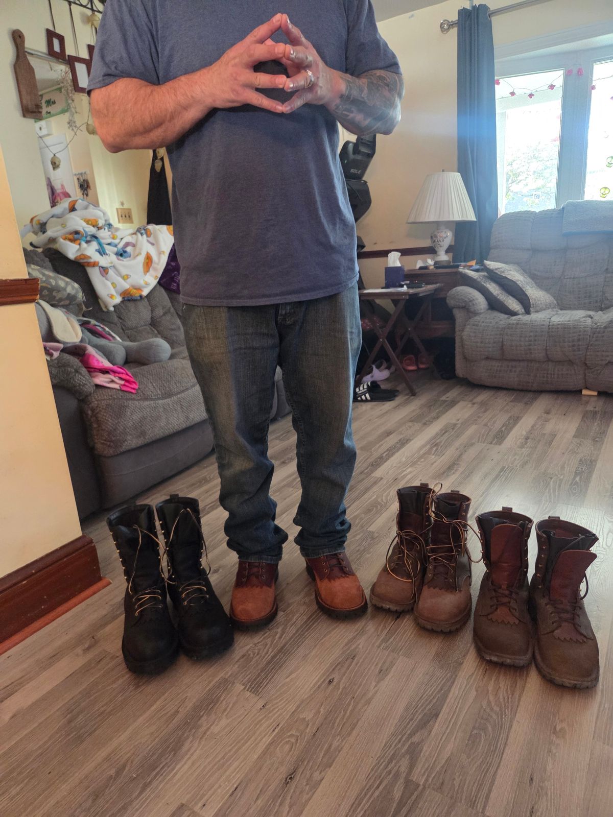 A person standing in a living room with several pairs of boots arranged on the wooden floor in front of them