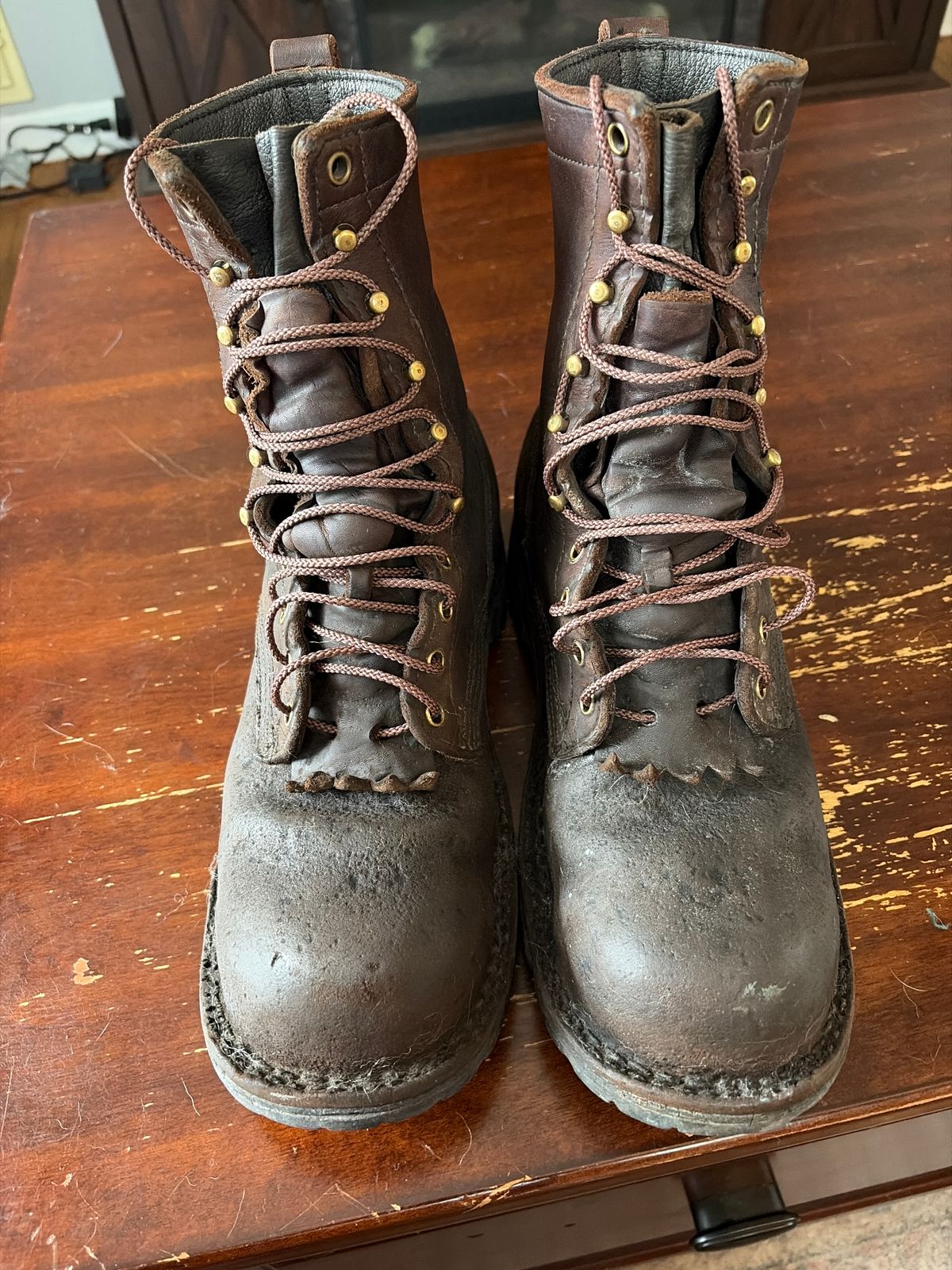 A pair of well-worn brown leather work boots with laces sitting on a wooden surface