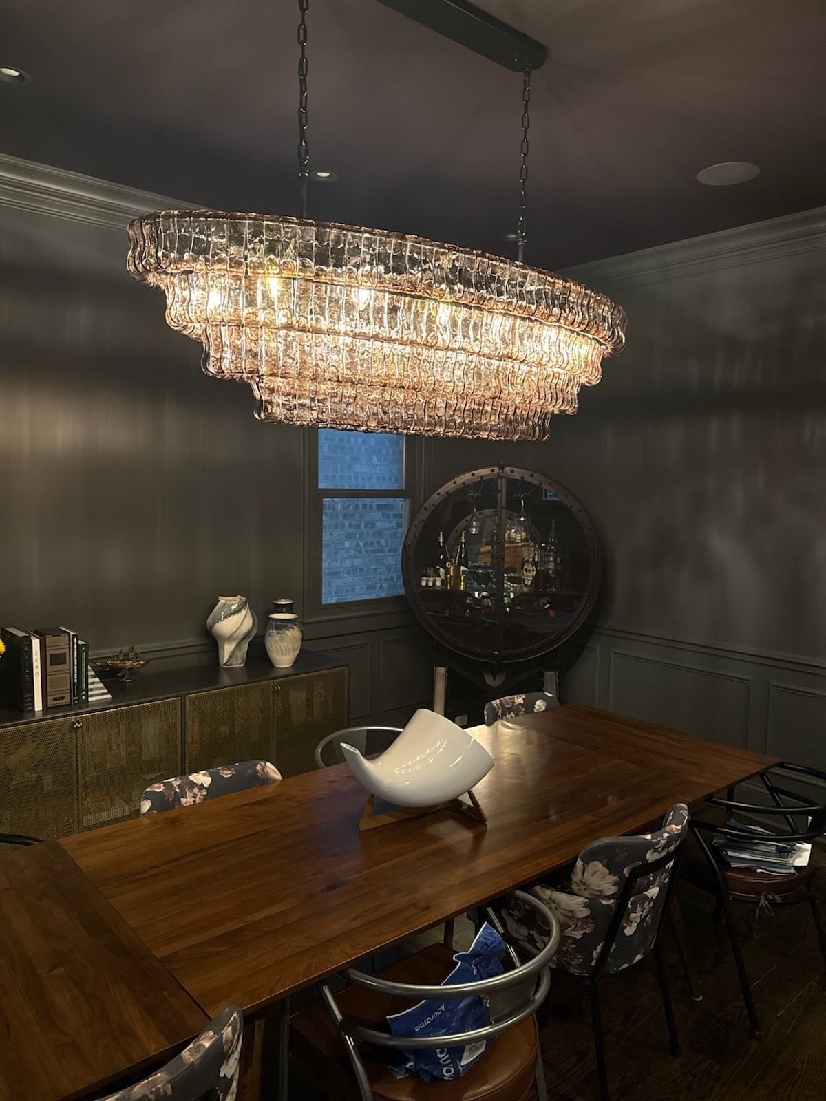 A modern dining room with dark walls, featuring a large crystal chandelier hanging over a wooden dining table with contemporary chairs, and a sideboard with decorative items against the wall.