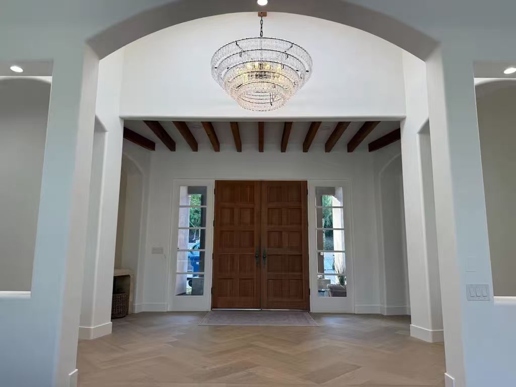 A modern foyer with white walls, wooden beams on the ceiling, a crystal chandelier, dark wooden double doors in the center, and sidelights on either side. The floor appears to be light-colored tile or stone.