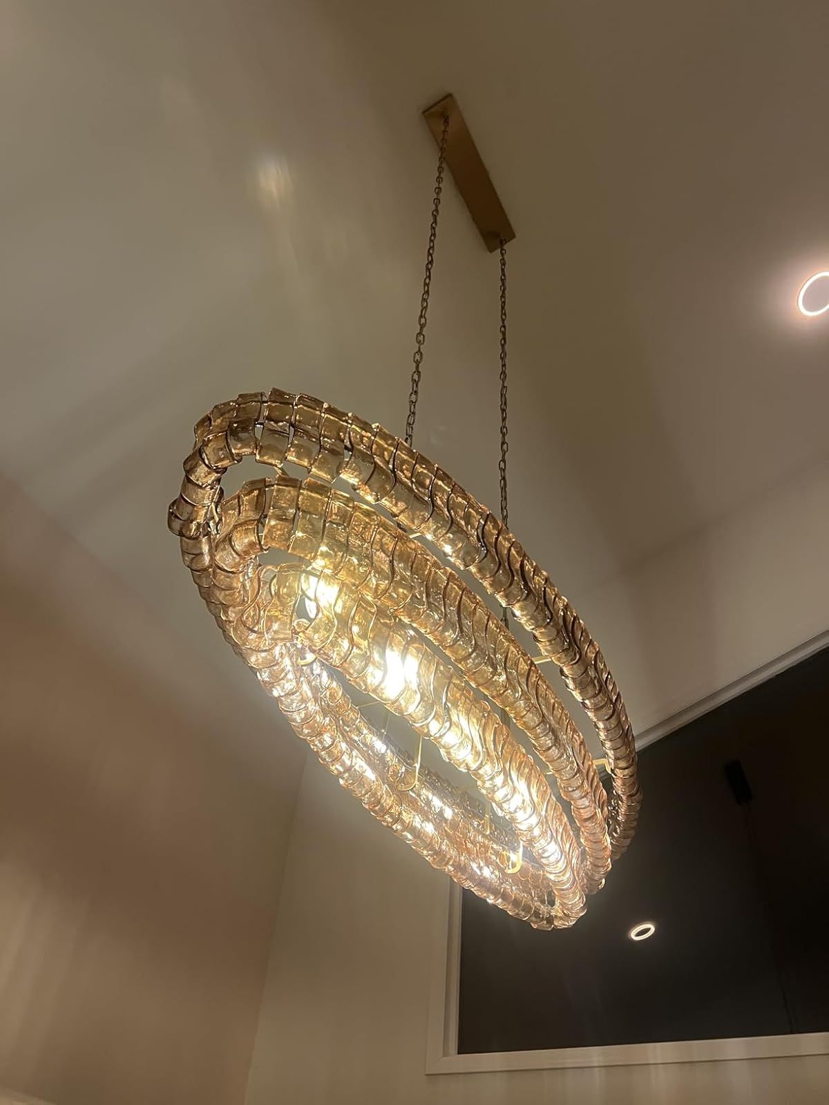 A modern pendant light fixture with a circular, layered glass design hanging from a brass chain and ceiling mount. The fixture emits warm golden light through its textured glass panels arranged in concentric rings.