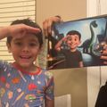 A young boy in pajamas poses with his hand to his forehead, mimicking the same gesture as an animated character on a book or poster behind him that features a nighttime city scene with dinosaurs.