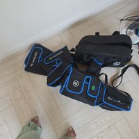 Customer-uploaded image. Click to see more details. The image represents: Recovery equipment laid out on wooden floor including black compression gear with blue trim and control unit, with person's feet visible wearing green striped pants