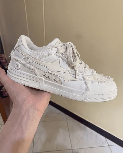 A hand holding a white sneaker with distressed details and rope-style laces against a neutral background
