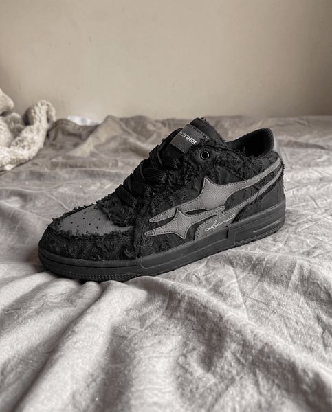 A black sneaker with textured fabric detailing sits on rumpled bedding in a bedroom setting