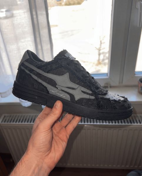 A hand holding a black and gray sneaker with a distressed, worn appearance near a window