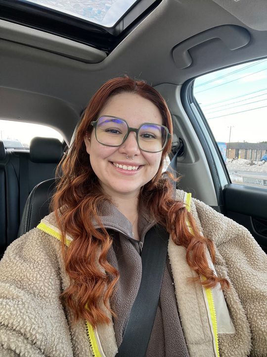 A woman with long reddish-brown hair and glasses smiling while sitting in the driver's seat of a car, wearing a beige and brown fleece jacket with yellow trim.