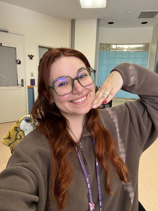 A smiling woman with long reddish-brown hair wearing glasses and a gray fleece jacket, posing with her hand near her face in what appears to be an office or classroom setting