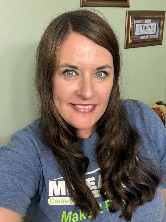 Woman with long brown hair wearing a blue t-shirt, smiling at camera in indoor setting with framed pictures on wall behind her
