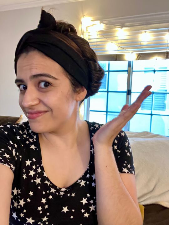 Woman with headband and star-patterned shirt waving at camera in bright room with window
