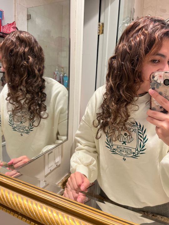 Person with curly hair taking a mirror selfie in a bathroom while wearing a cream-colored sweatshirt