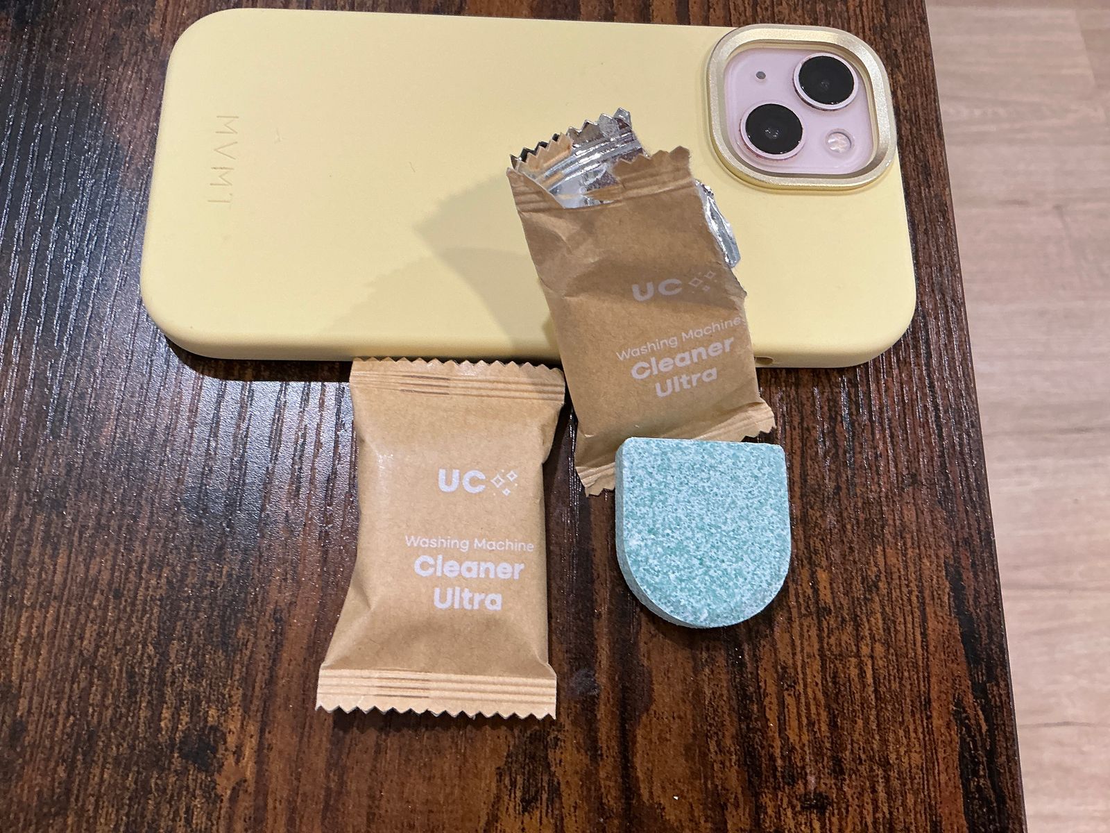 A yellow phone case next to two brown packets labeled "UC Washing Machine Cleaner Ultra" and a blue cleaning tablet on a wooden surface