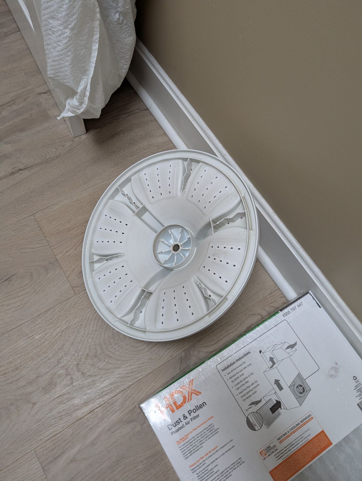 White circular washing machine drum or basket with perforated holes sitting on wooden floor next to instruction manual