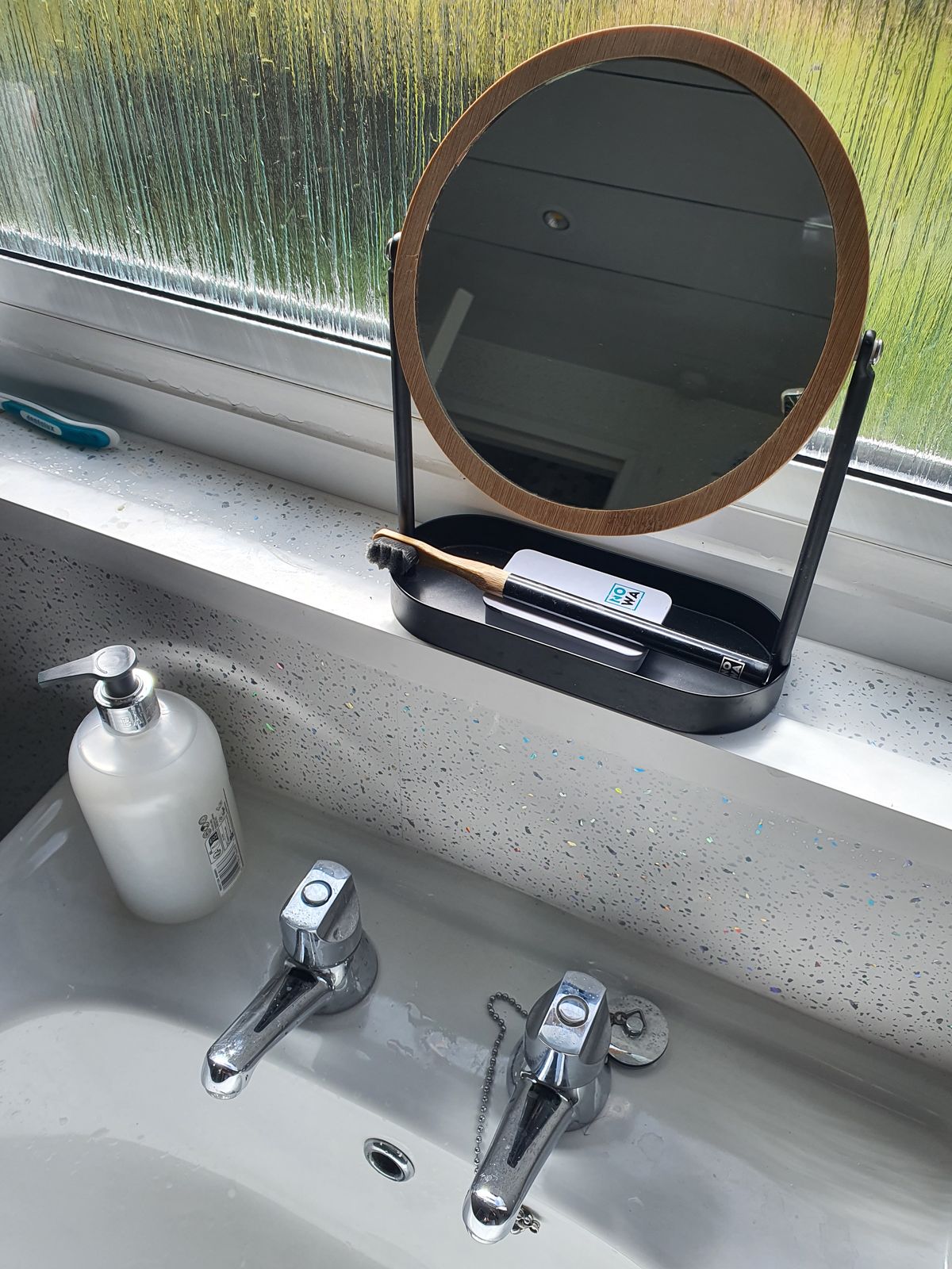 A bathroom sink with chrome faucets, a pump bottle of soap, and a round mirror with wooden frame sitting on the windowsill