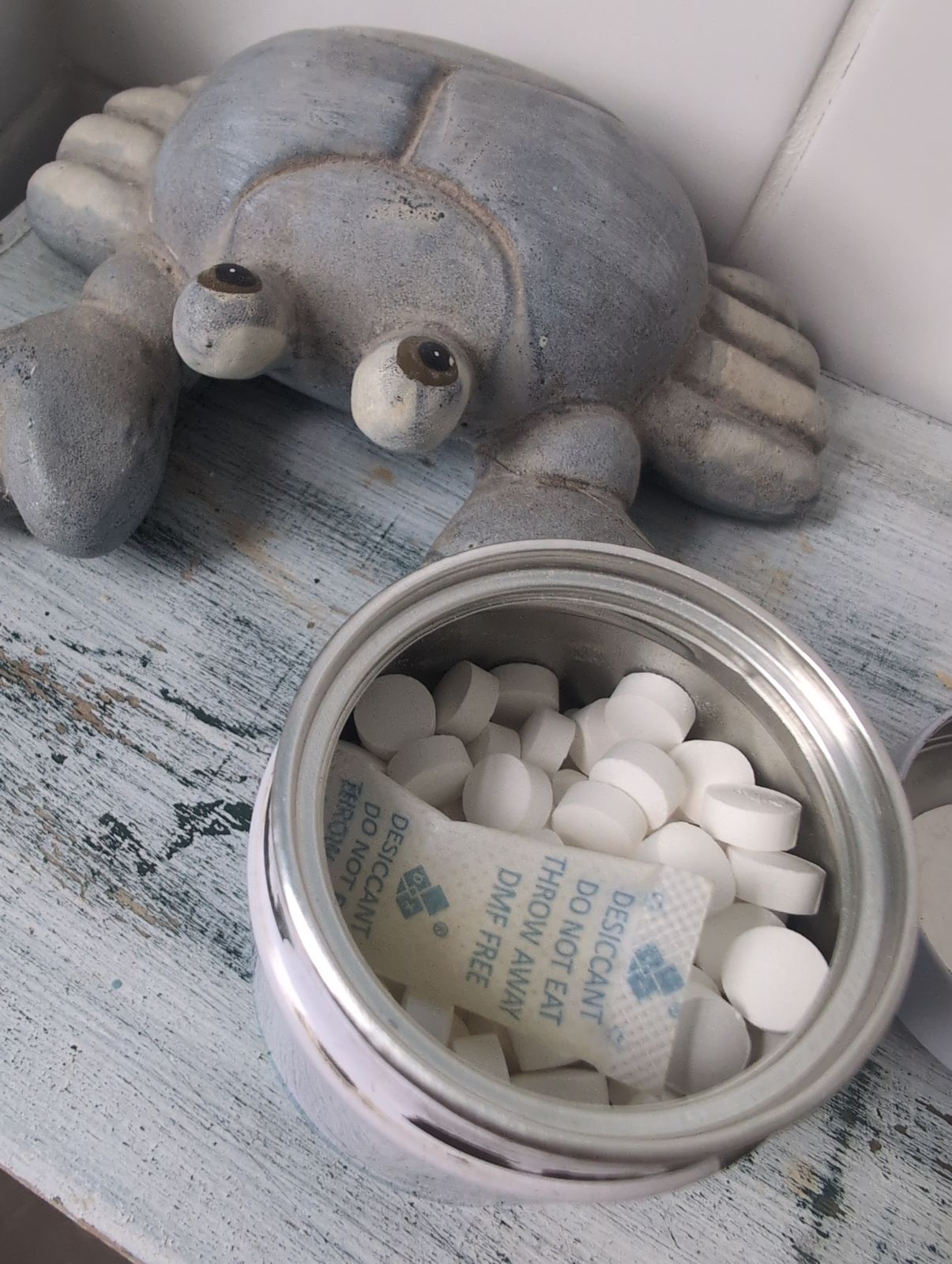 A ceramic crab figurine sits next to an open metal container filled with white tablets and a small packet of desiccant on a weathered wooden surface.