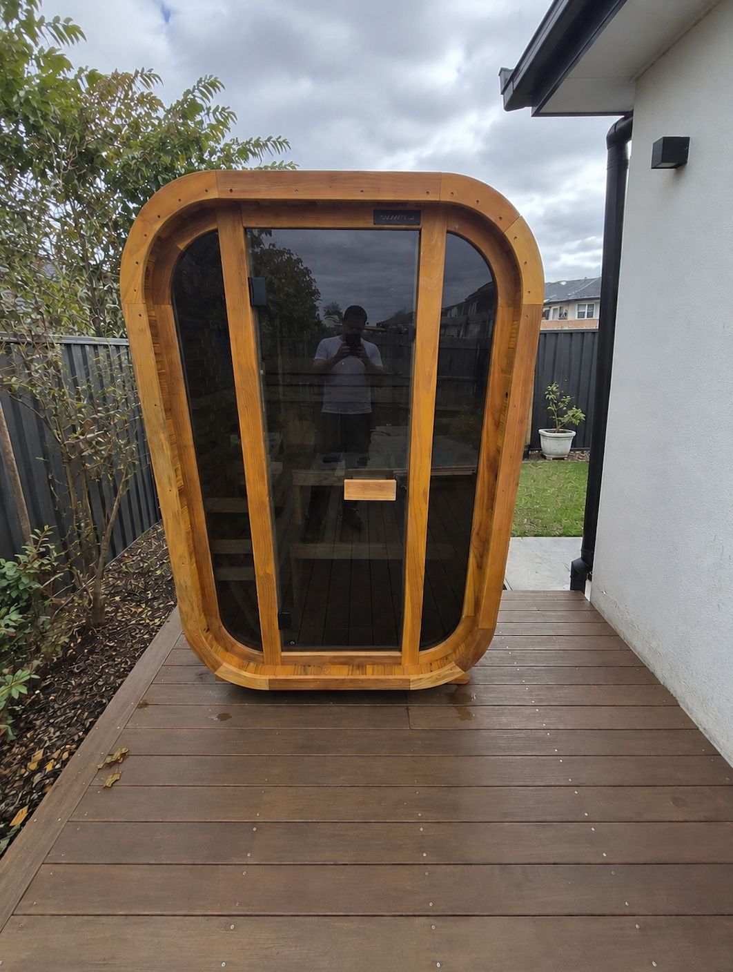 A wooden pod-like structure with curved walls and dark tinted windows sits on a brown wooden deck in a residential backyard. A person is visible inside the pod. The setting includes a white house wall, fencing, landscaping with plants, and an overcast sky.