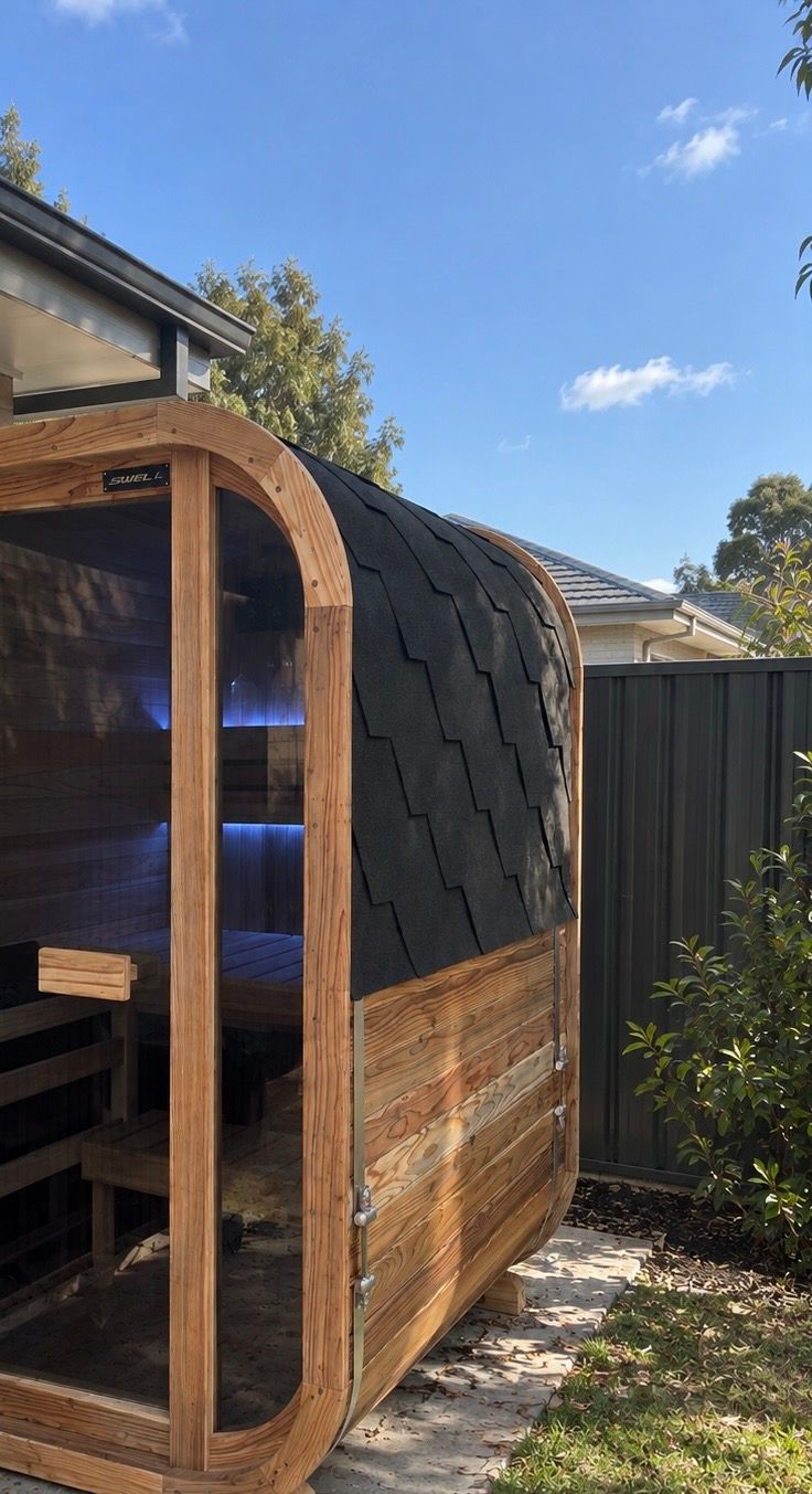 A modern wooden sauna pod with curved architecture, featuring a wooden frame, dark shingle roof, and glass door with blue interior lighting, situated in a residential backyard on a sunny day.