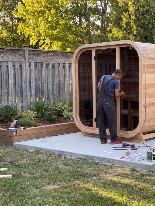 A worker in dark clothing installs or assembles a wooden sauna pod in a backyard, standing on a concrete pad next to a raised garden bed with plants and trees in the background.
