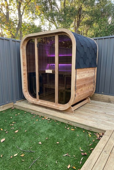 A modern wooden sauna pod with curved architectural design, featuring large glass doors and purple interior lighting, positioned on a wooden deck in a residential backyard surrounded by metal fencing and trees.
