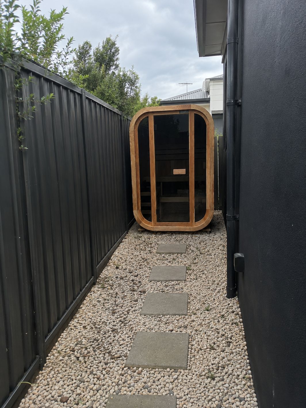 A wooden sauna cabin with rounded corners and dark glass door sits in a narrow gravel pathway between two black metal fences, with stepping stones leading to it and green foliage visible above the fences.