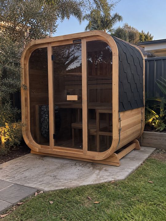 A modern wooden sauna cabin with curved corners and large glass doors, situated in a backyard garden setting on a concrete pad. The structure features natural wood framing, black shingled roof panels, and interior wooden benches visible through the transparent front panels. Surrounding landscaping includes palm trees, olive trees, and manicured grass.