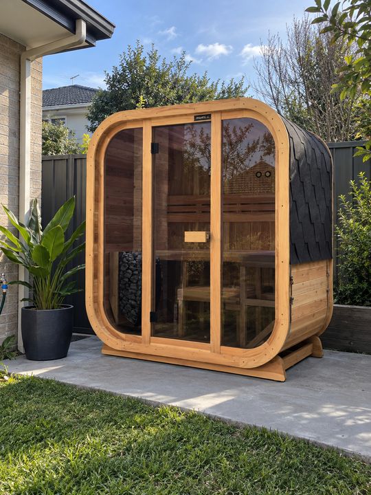A modern wooden sauna pod with curved glass doors and windows, positioned on a patio in a residential backyard. The sauna features natural wood framing with a dark roof, and is surrounded by landscaping including potted plants and green hedges.
